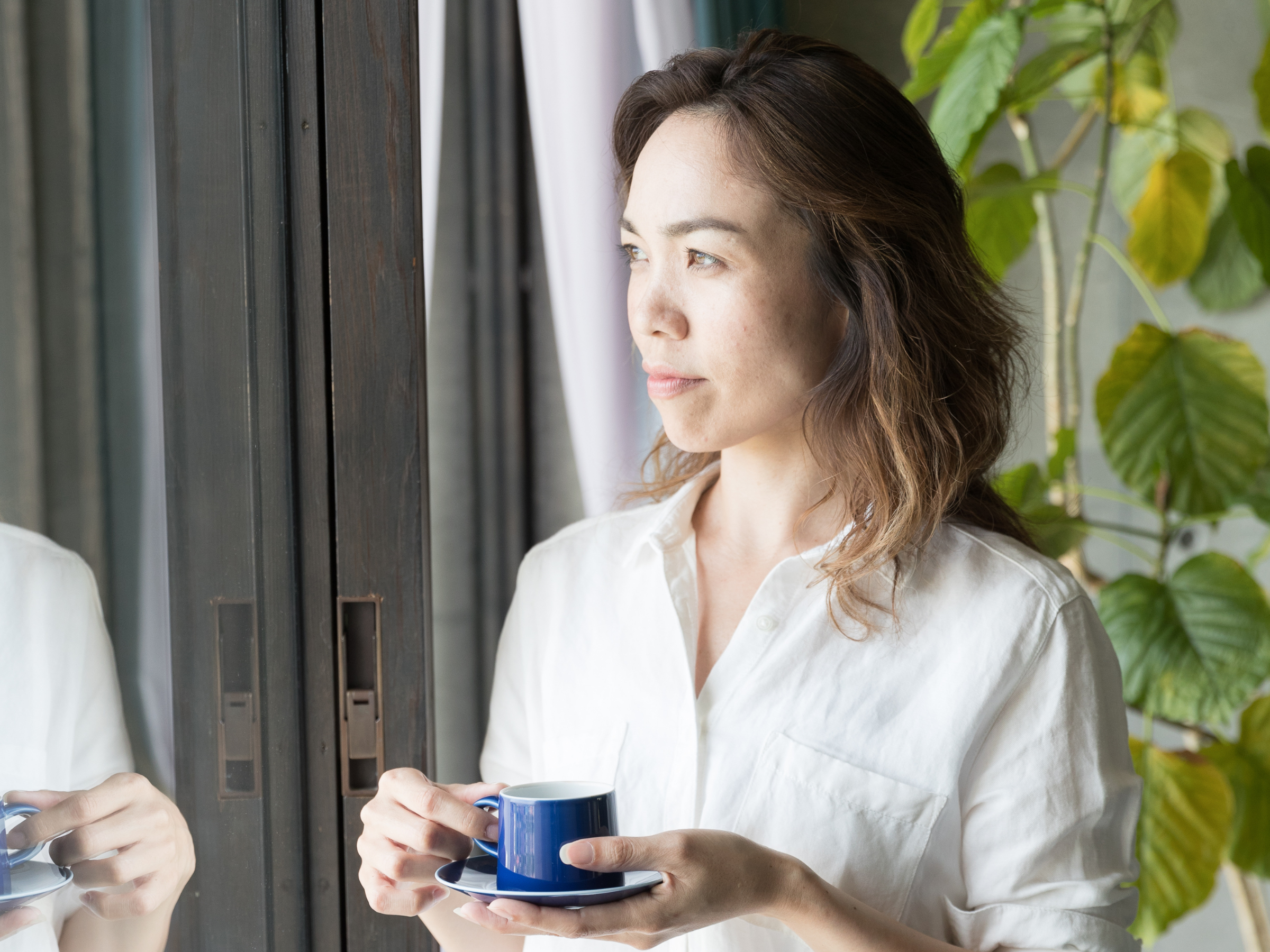 Woman looking out the glass window in a reflective manner, holding a cup of hot drink in her hand